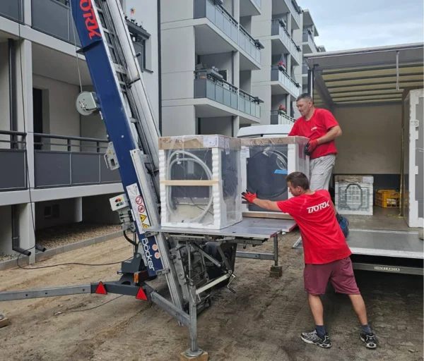 Zwei Bork-Mitarbeiter in roten T-Shirts laden eine verpackte Waschmaschine auf einen Möbellift vor einem Mehrfamilienhaus