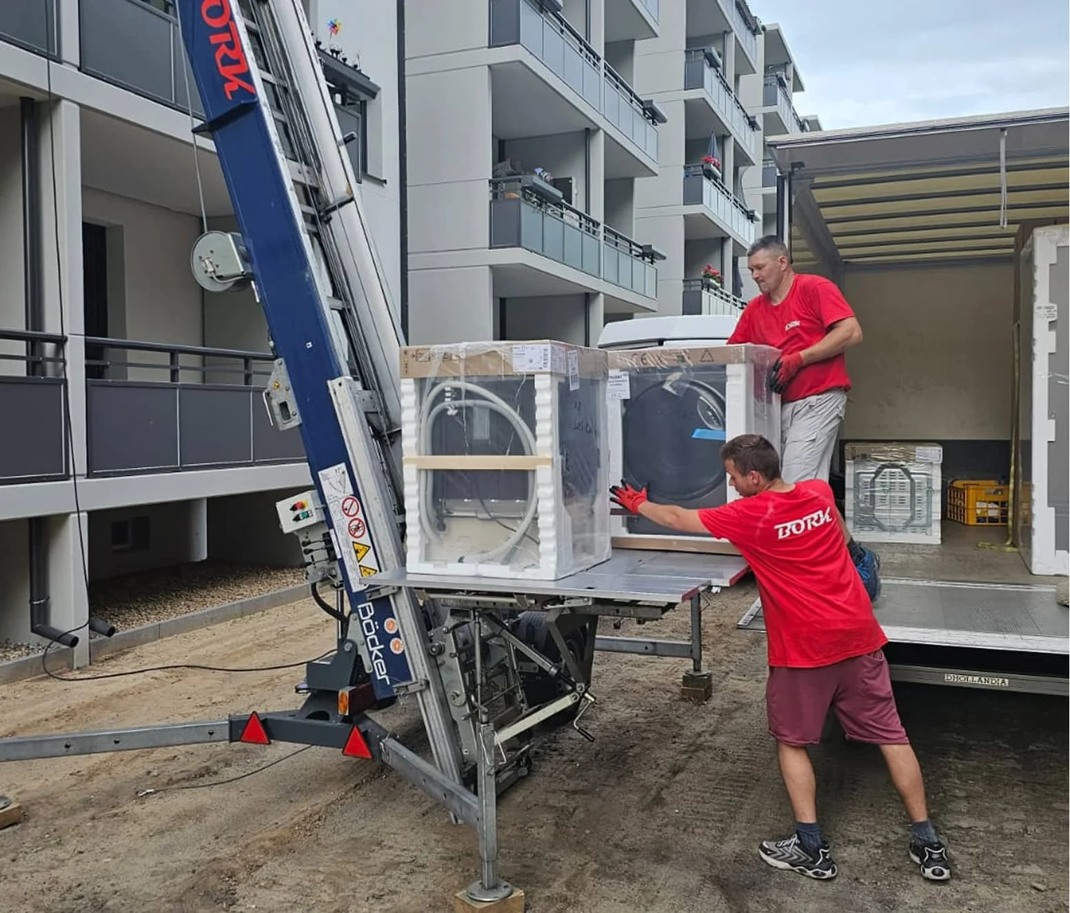 Zwei Bork-Mitarbeiter in roten T-Shirts laden eine verpackte Waschmaschine auf einen Möbellift vor einem Mehrfamilienhaus