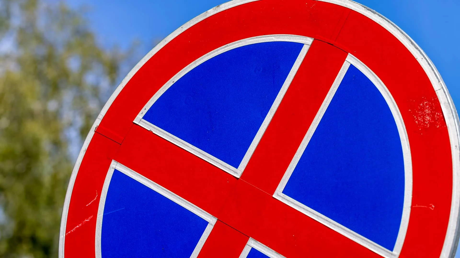 Nahaufnahme eines deutschen Halteverbotszeichens (blaues Rundschild mit rotem Kreuz) vor blauem Himmel