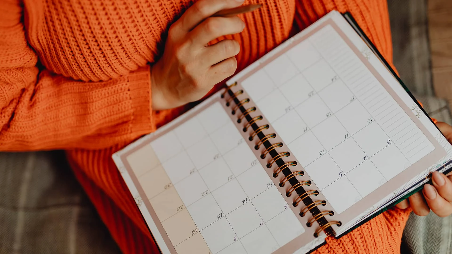 Person im orangefarbenen Pullover hält einen aufgeschlagenen Kalender-Terminplaner